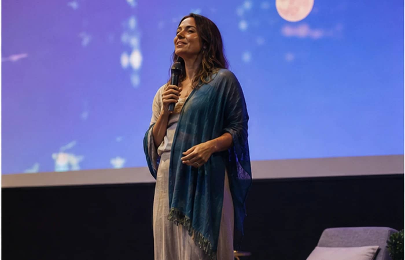 Mulher falando em palco com projeção azul ao fundo.